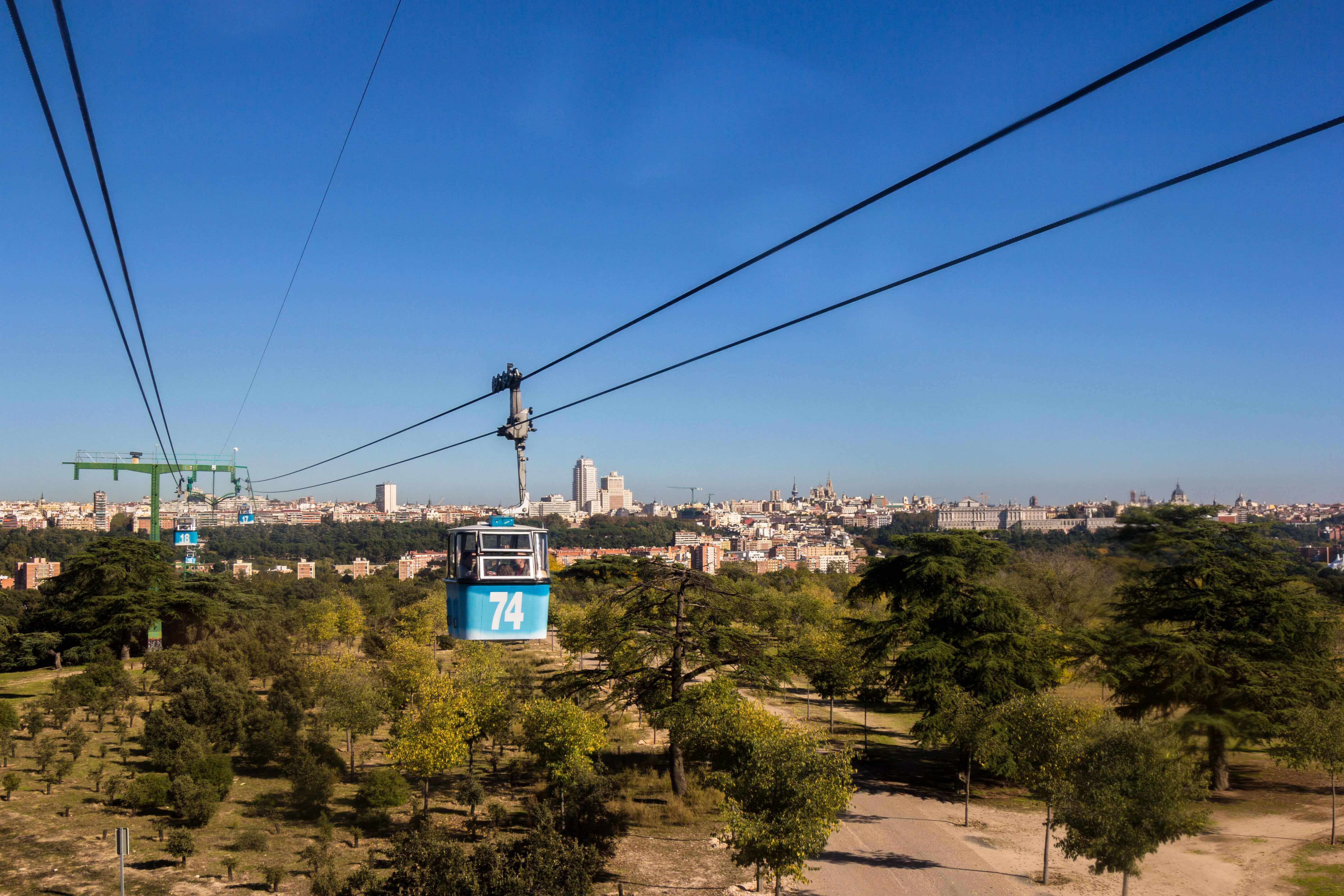 A blue cable car runs across a beautiful park. In the background the skyline of a city can be seen. Casa de Campo, Madrid, Spain.